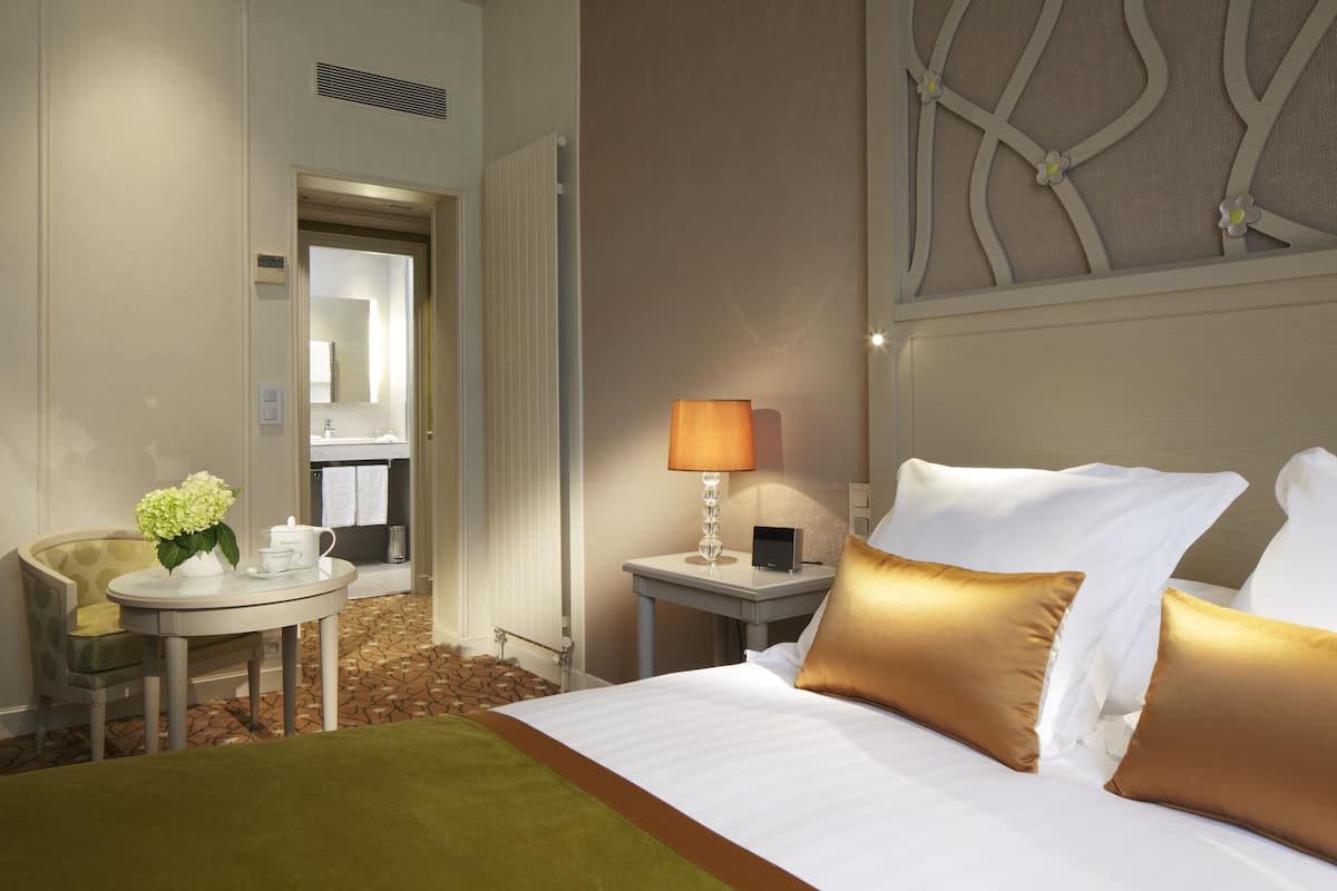 A hotel room with a bed, a bedside table with a lamp, a chair, and a small table with a vase of flowers.