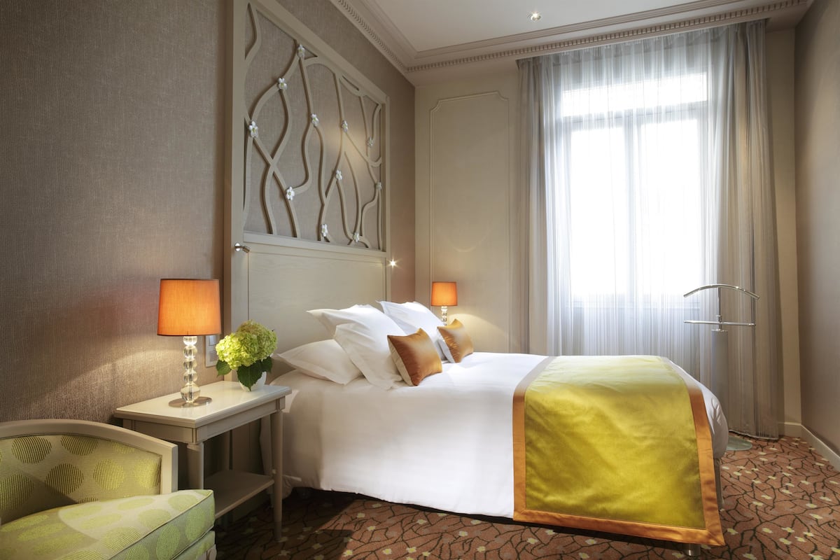 A hotel room with a large bed, two bedside lamps, a nightstand with a lamp, a chair, and a decorative wall panel.