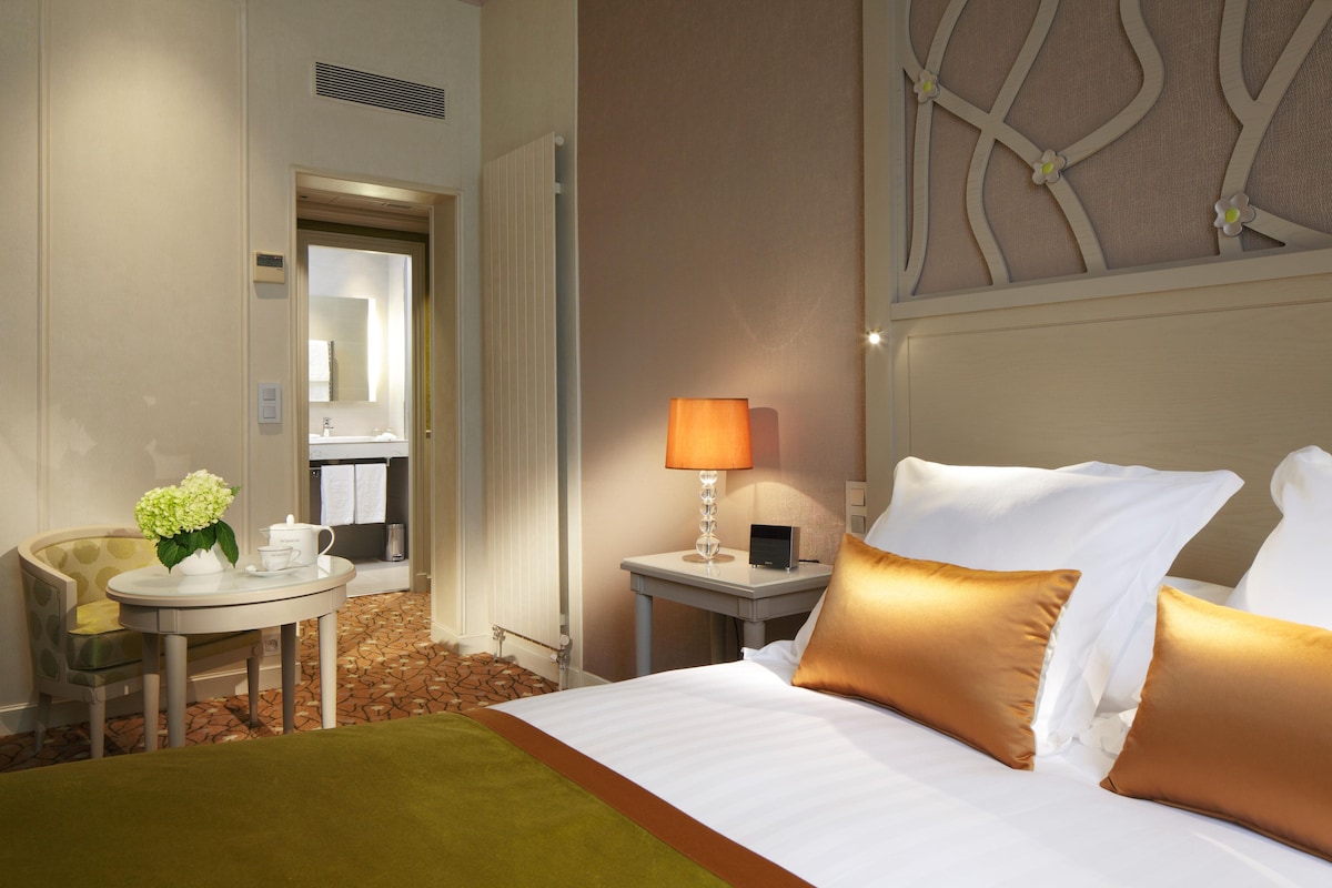A hotel room with a bed, a bedside table with a lamp, a chair, and a small table with a vase of flowers.