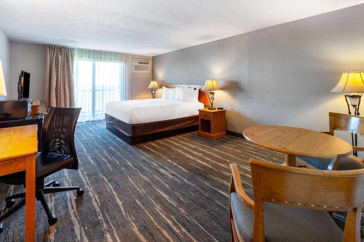 Hotellihuone, jossa on sänky, kaksi yöpöytää valaisimineen, työpöytä tuolilla sekä pieni pyöreä pöytä.