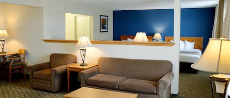 Habitación de hotel con sofá, sillón y una pequeña mesa. Cuenta con una cama con cabecera y una mesita de noche con lámpara.