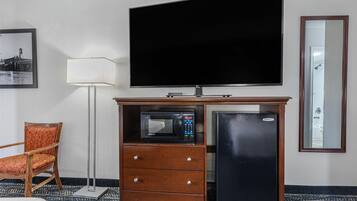 A hotel room with a flat-screen TV, a microwave, a refrigerator, a wooden cabinet, a chair, and a floor lamp.