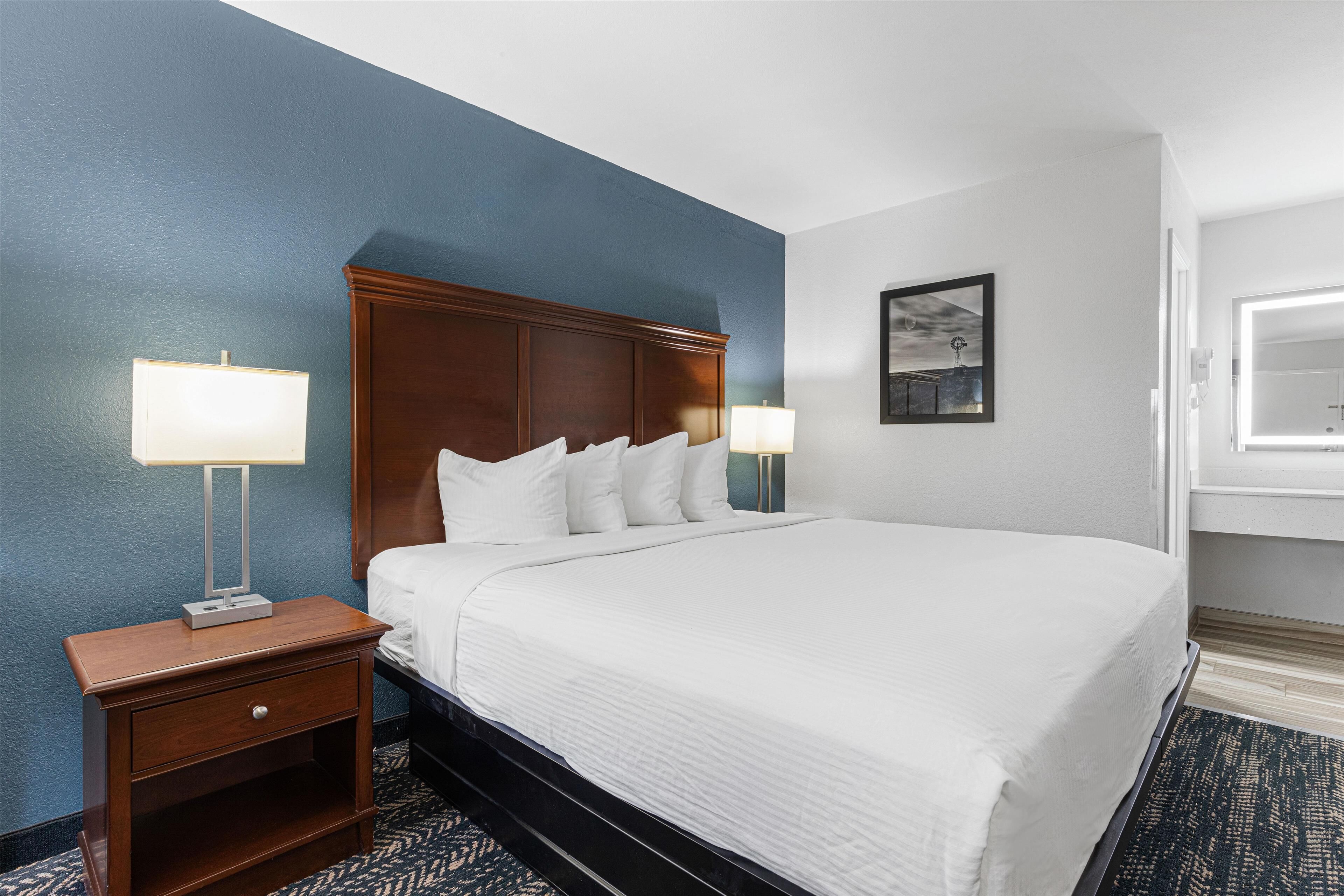 Una habitación de hotel con una cama grande, dos mesitas de noche con lámparas, un pequeño mueble auxiliar y un cuadro en la pared, además de una ventana con vistas.
