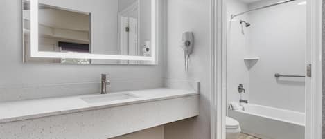 Un baño moderno con un espejo grande, un lavabo blanco, un secador de pelo montado en la pared y una zona de ducha.
