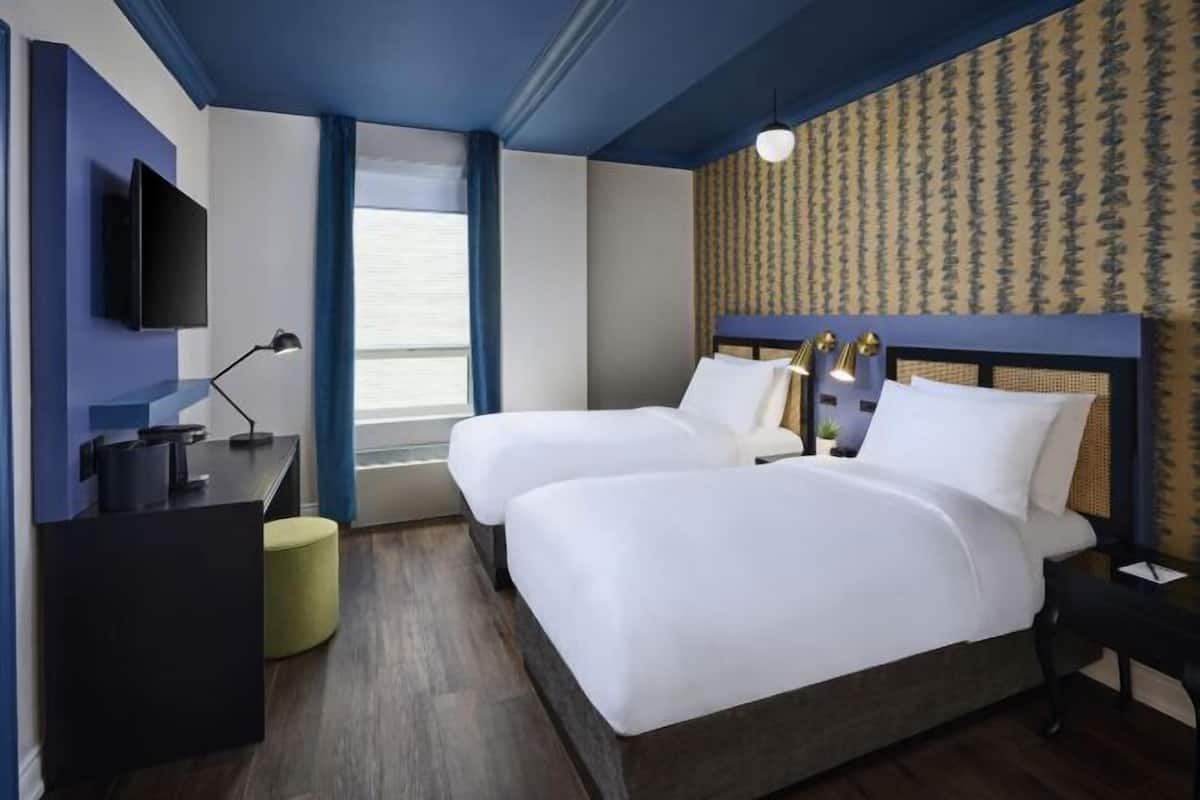 A hotel room with two beds, a desk, a TV, and a window with curtains.