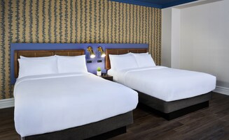 Hotellihuone, jossa on kaksi sänkyä, puinen pääty, yöpöytä lampun kanssa ja kuviollinen tapetti.