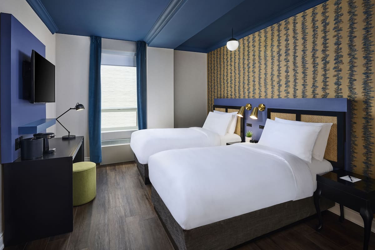 A hotel room with two beds, a flat-screen TV, a desk with a lamp, and a window with curtains.