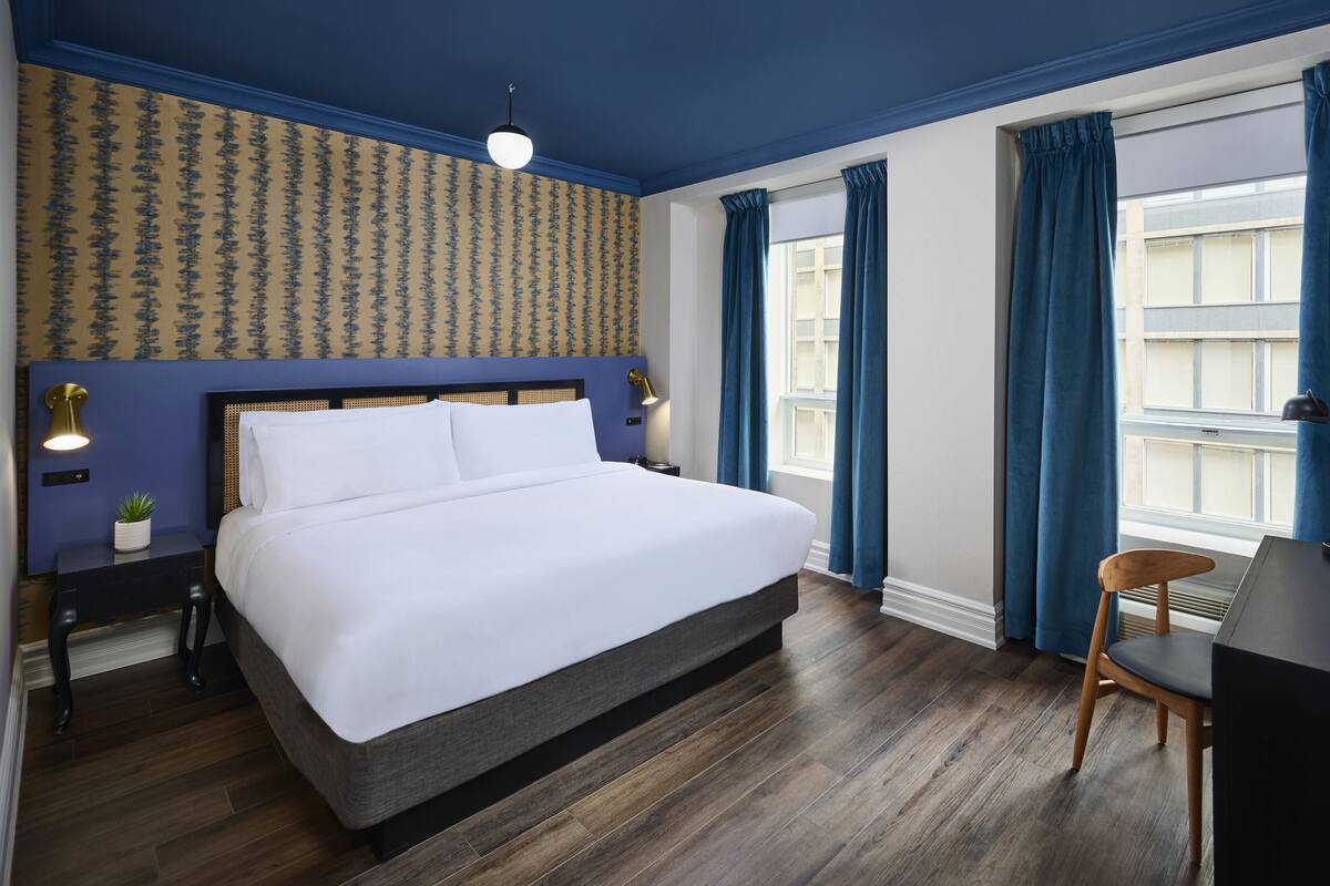 A neatly arranged hotel room with a large bed, a desk with a chair, a desk lamp, a small plant, and a window with curtains.