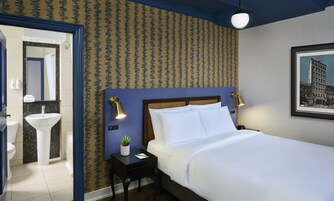 Een hotelkamer met een bed, nachtkastje, lamp en een badkamer met wastafel en spiegel.