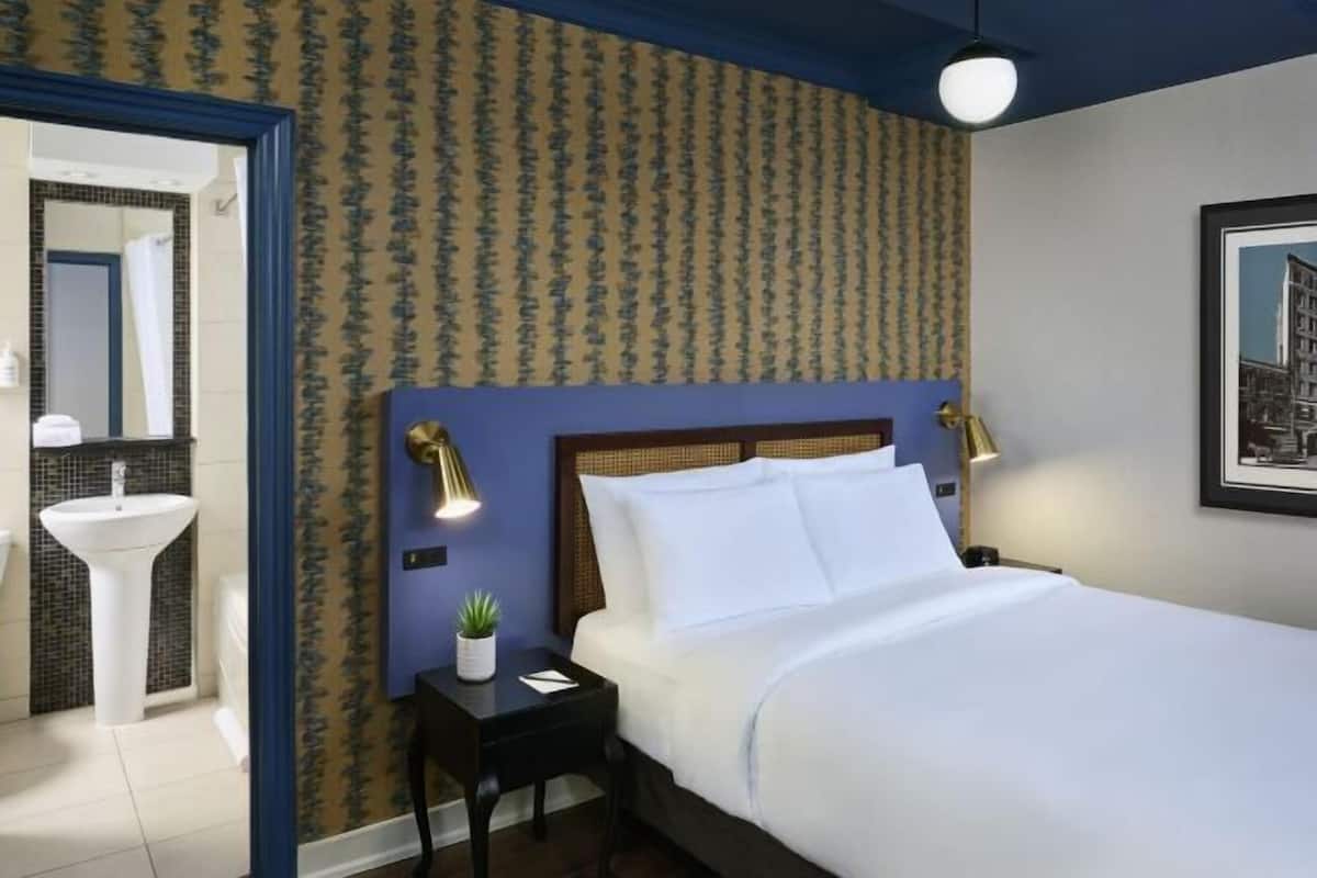 A hotel room with a bed, bedside lamps, a small table with a plant, a mirror, and a bathroom with a sink and shower.