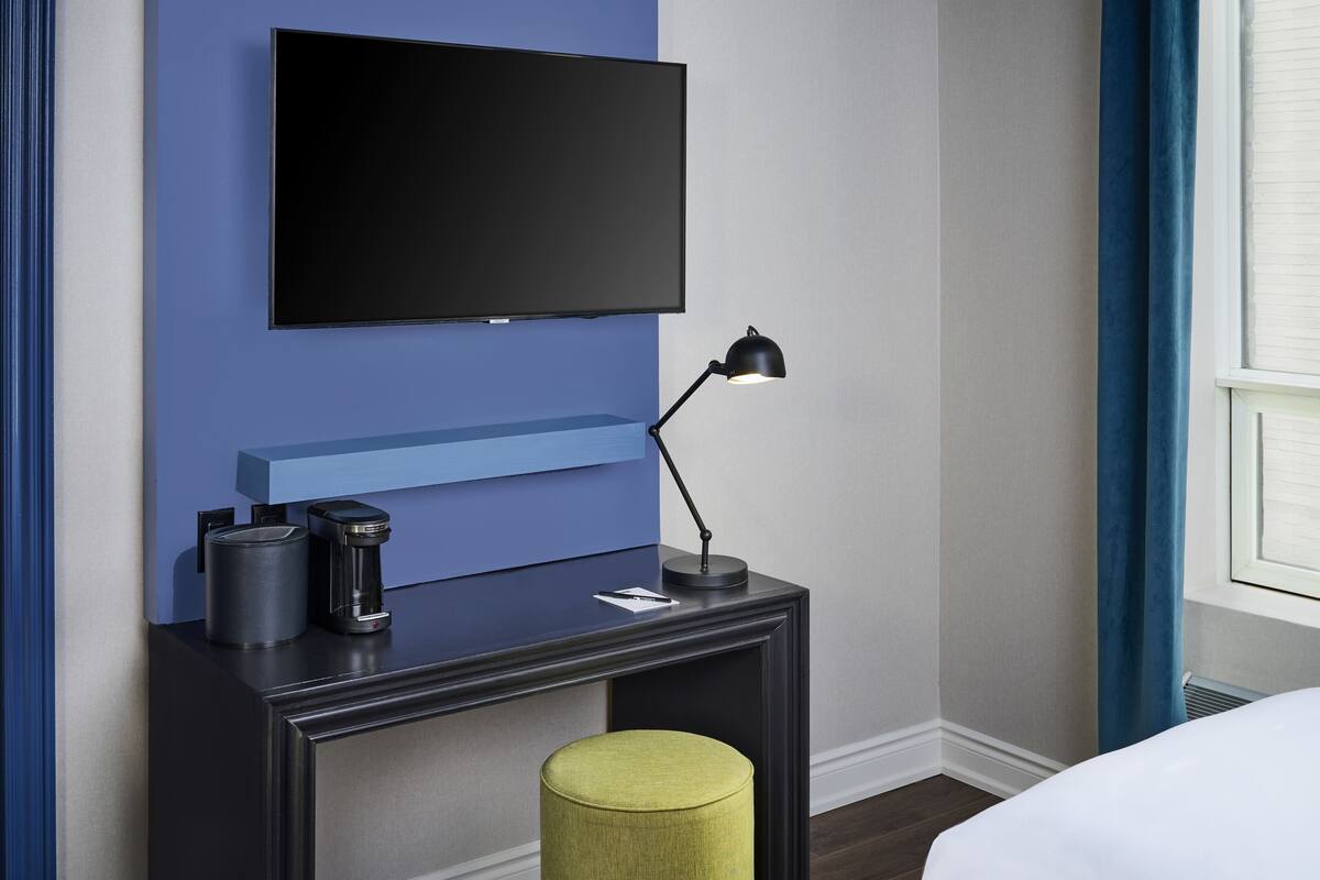 A modern hotel room with a flat-screen TV mounted on a blue wall, a small bedside table with a lamp, a coffee maker, and a green ottoman.