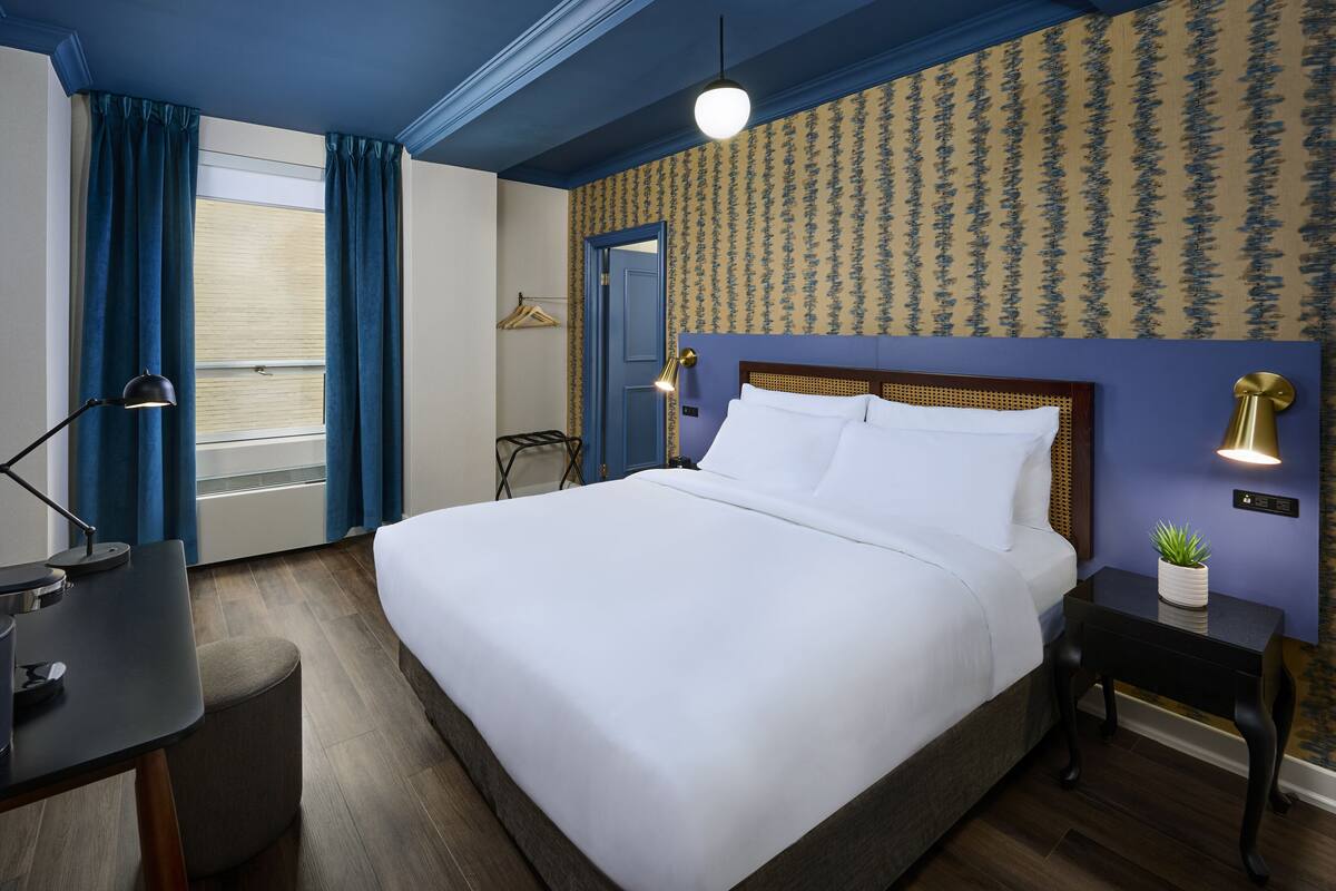 A hotel room with a large bed, a desk with a lamp, a small table with a plant, and a wall-mounted lamp.