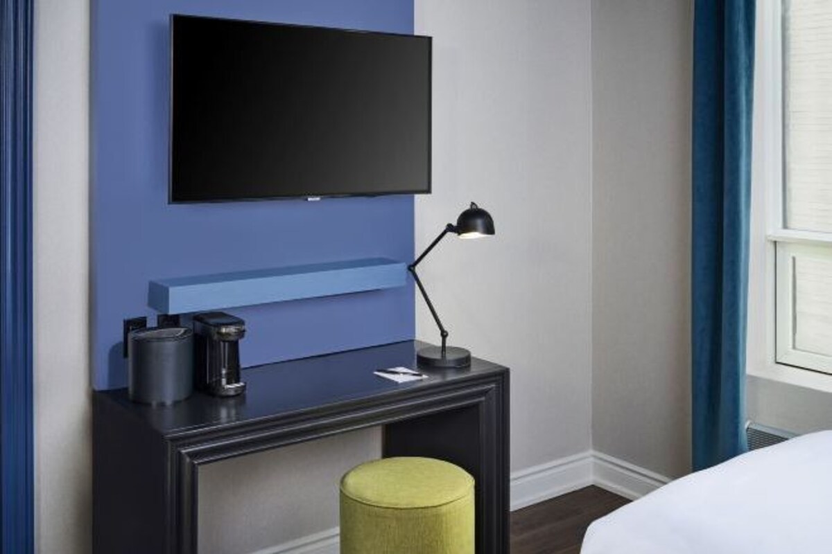 A hotel room with a flat-screen TV mounted on a blue wall, a bedside table with a lamp, a coffee maker, and a yellow ottoman.