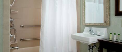 Baño con bañera, ducha, cortina blanca, lavabo, espejo y barras de apoyo.