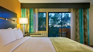 A hotel room with a bed, a nightstand with a lamp, a window with curtains, and a view of trees outside.