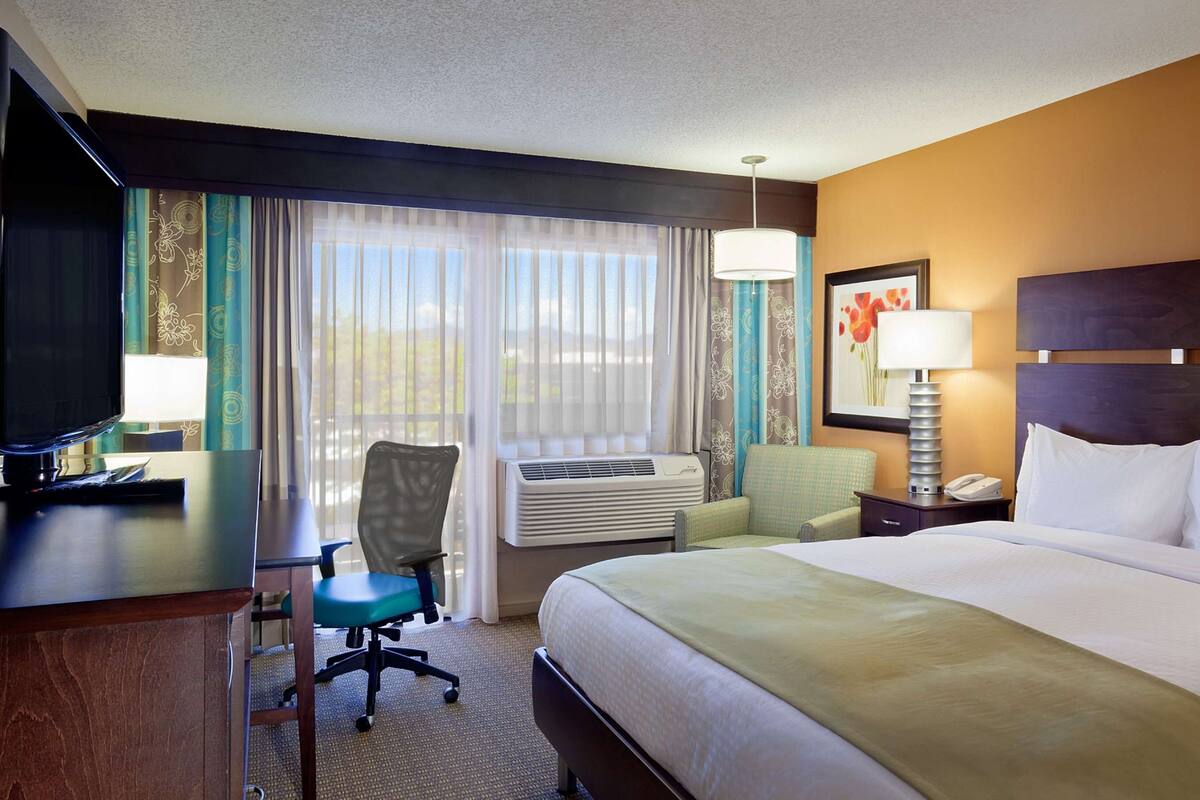 A hotel room with a bed, desk, chair, air conditioning unit, and a television.