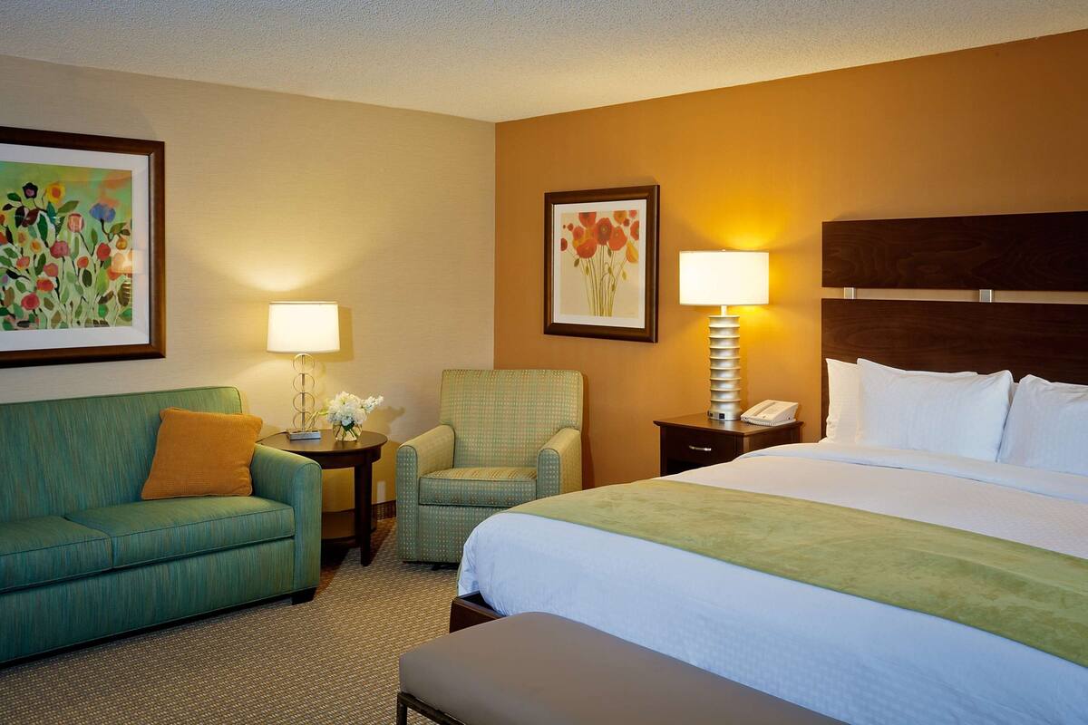 A hotel room with a bed, a green armchair, a sofa, a nightstand with a lamp, and two framed floral paintings.