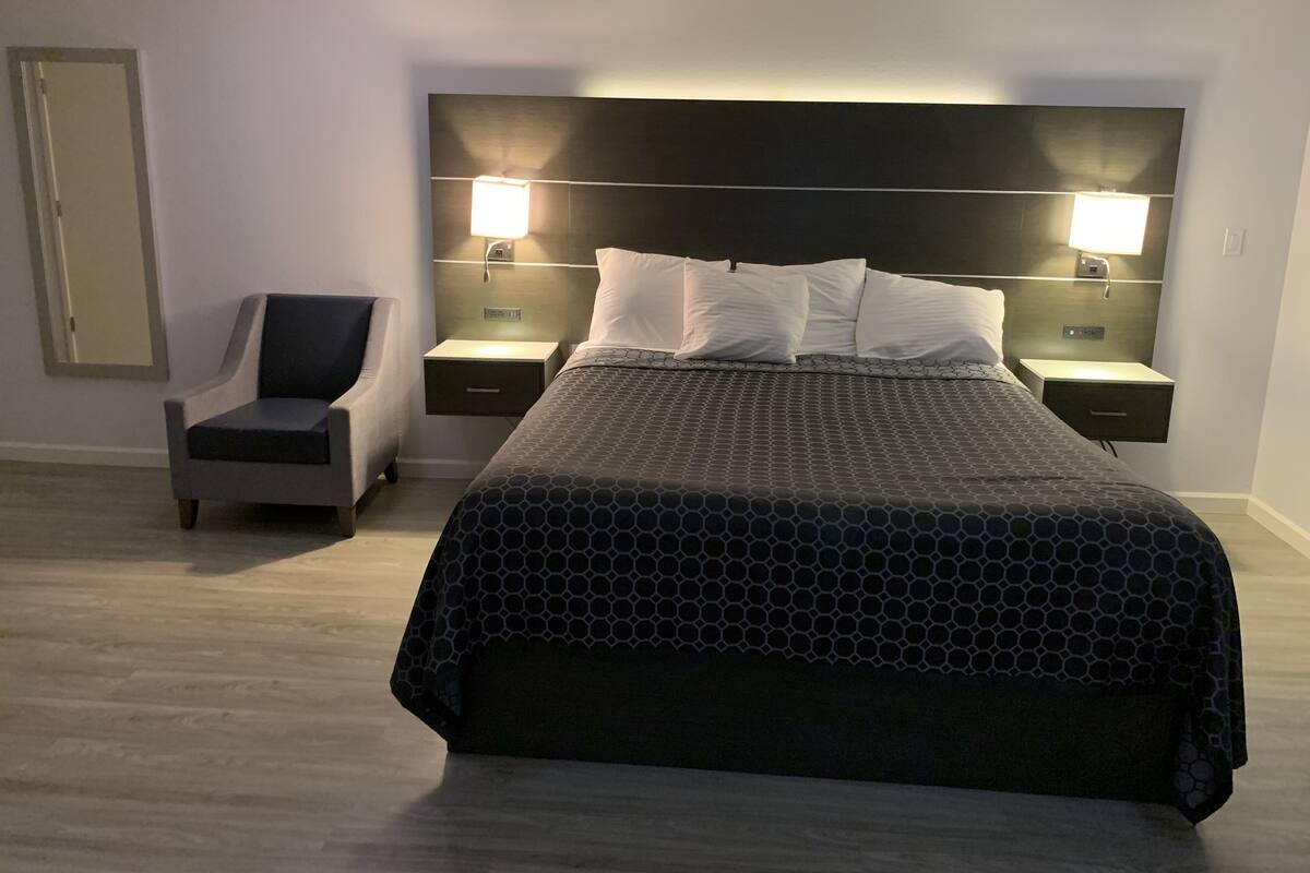 A modern hotel room with a large bed, two bedside tables with lamps, a chair, and a mirror on the wall.
