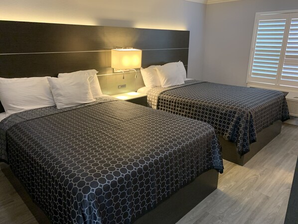 A hotel room with two beds, a nightstand with a lamp, and a window with blinds.
