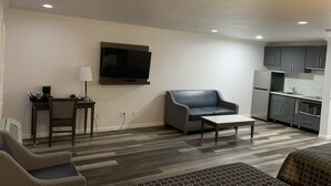 Een moderne woonkamer met een flatscreen televisie, een bank, een salontafel, en een keukenhoek met kasten en apparatuur.