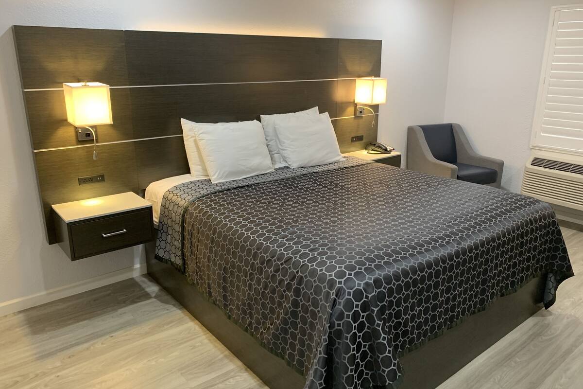 A modern hotel room with a large bed, two bedside lamps, a nightstand, a chair, and a built-in heater.