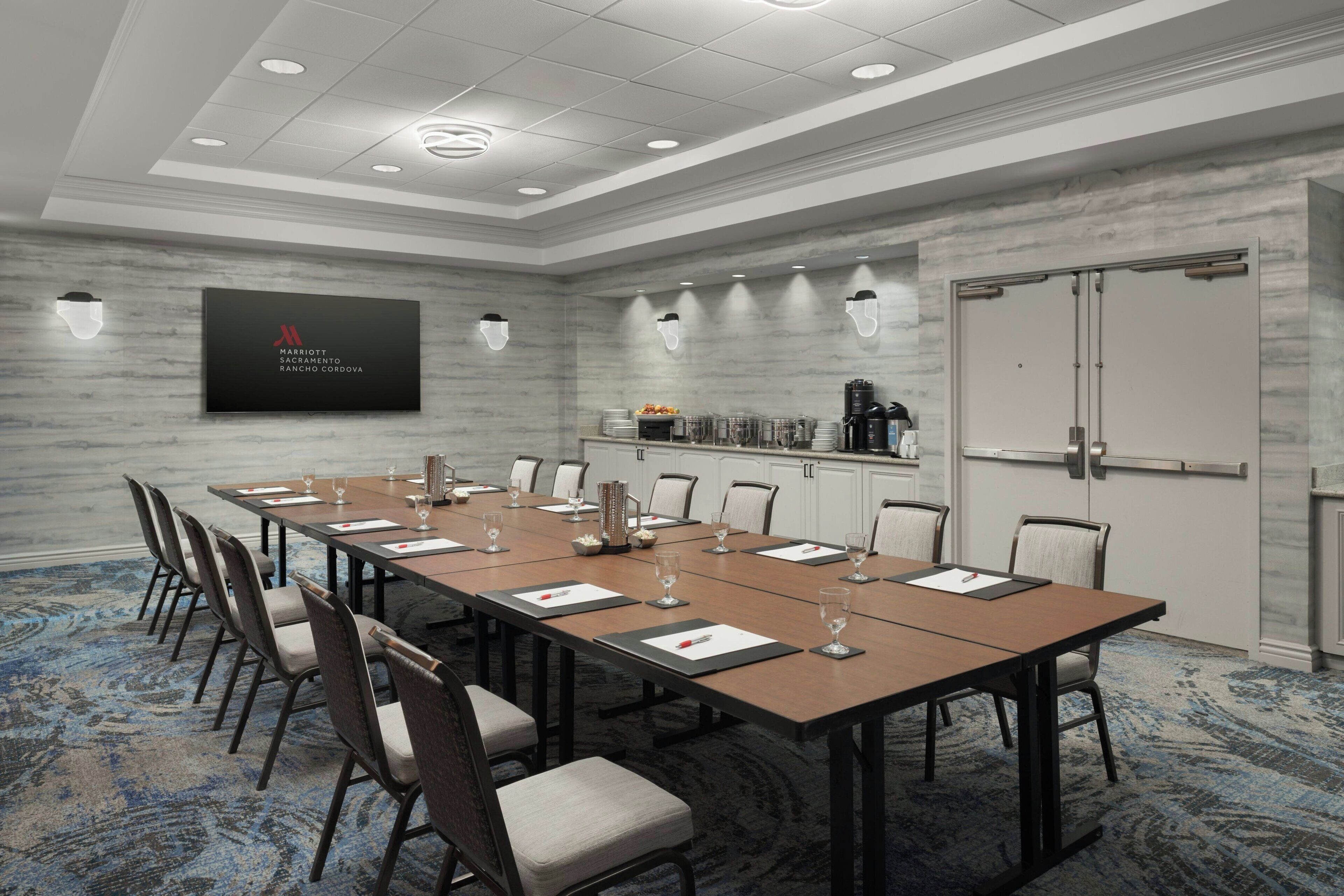 A conference room with a long table, chairs, a screen displaying the hotel name, and a kitchenette area.