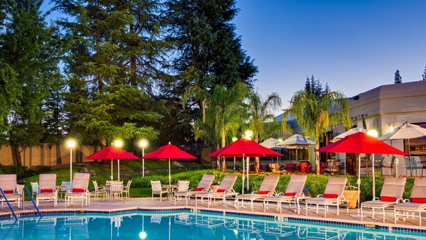 A pool area with lounge chairs and red umbrellas, surrounded by trees and a building in the background.