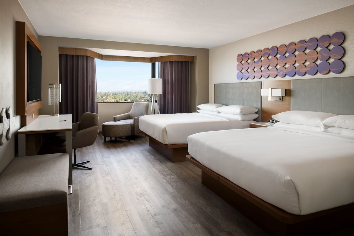 A hotel room with two beds, a desk, a chair, a TV, and a large window with curtains.