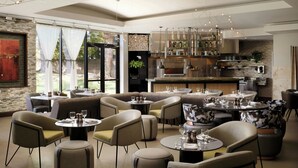 A modern restaurant with round tables and cushioned chairs, a bar area with bottles, and a view of the outdoors.