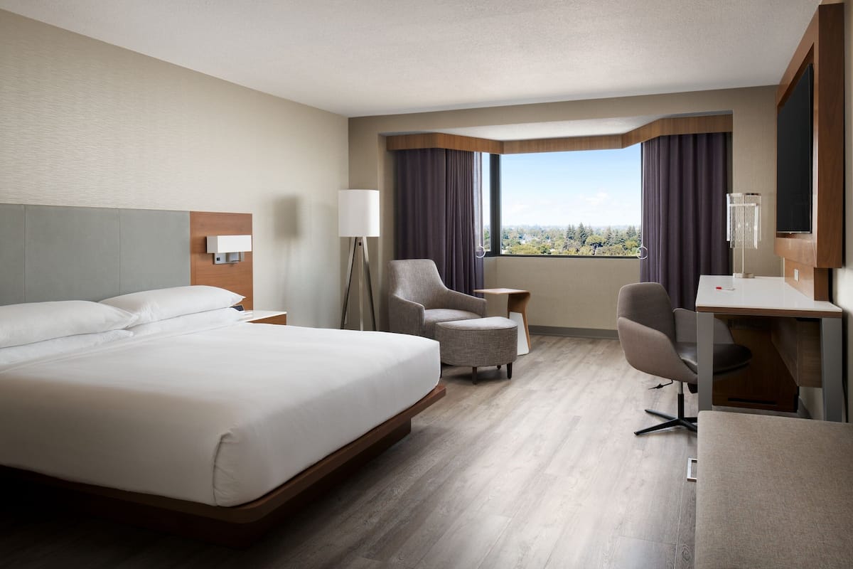 A hotel room with a large bed, a desk, two armchairs, a side table, and a view of the city.