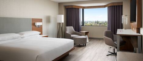 Una habitación de hotel con una cama grande, un escritorio, dos sillones, una mesita auxiliar y vistas de la ciudad.
