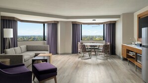 A modern hotel room with a sofa, armchair, ottoman, dining table with chairs, and a kitchen area.
