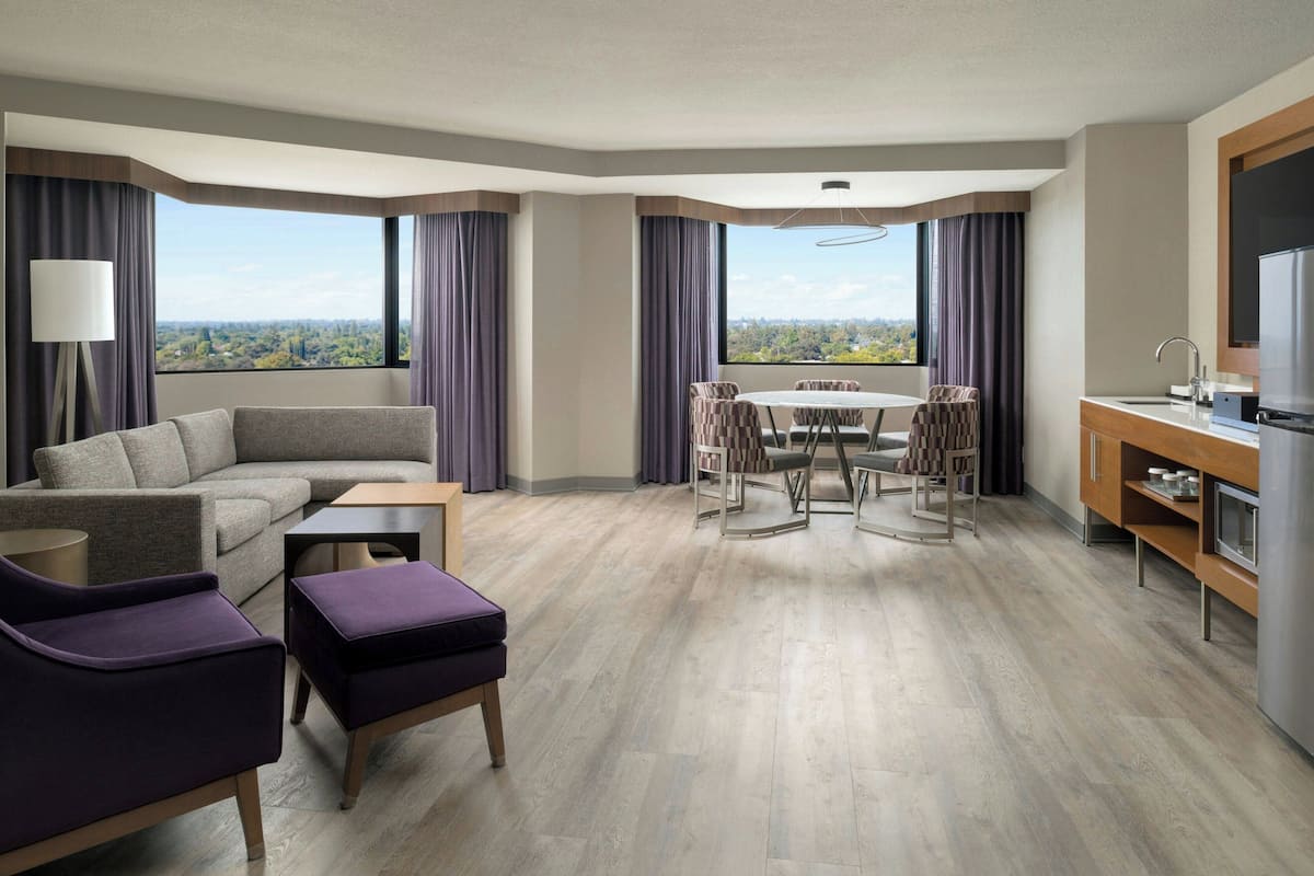 A modern hotel room with a sofa, armchair, ottoman, dining table with chairs, and a kitchen area.