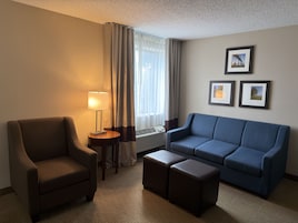 Quarto de hotel com uma poltrona marrom, um sofá azul, dois pufes, uma luminária, uma janela com cortinas e três quadros na parede.