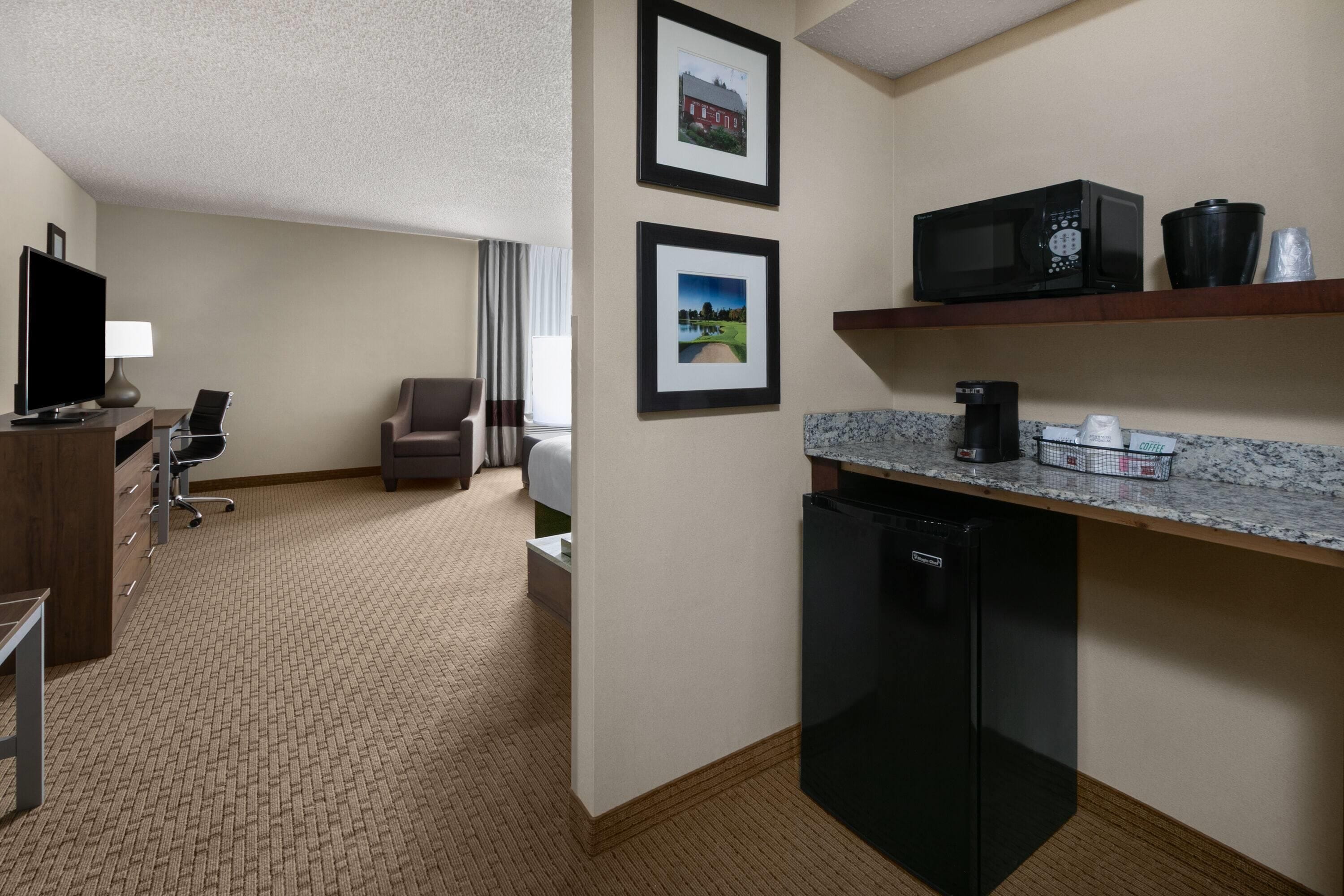 A hotel room with a kitchenette, a television, a desk, a chair, and two framed pictures on the wall.