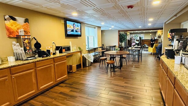 A spacious restaurant with wooden flooring, a central dining area, and a kitchen counter with various appliances. - Wingate by Wyndham Utica (Utica)