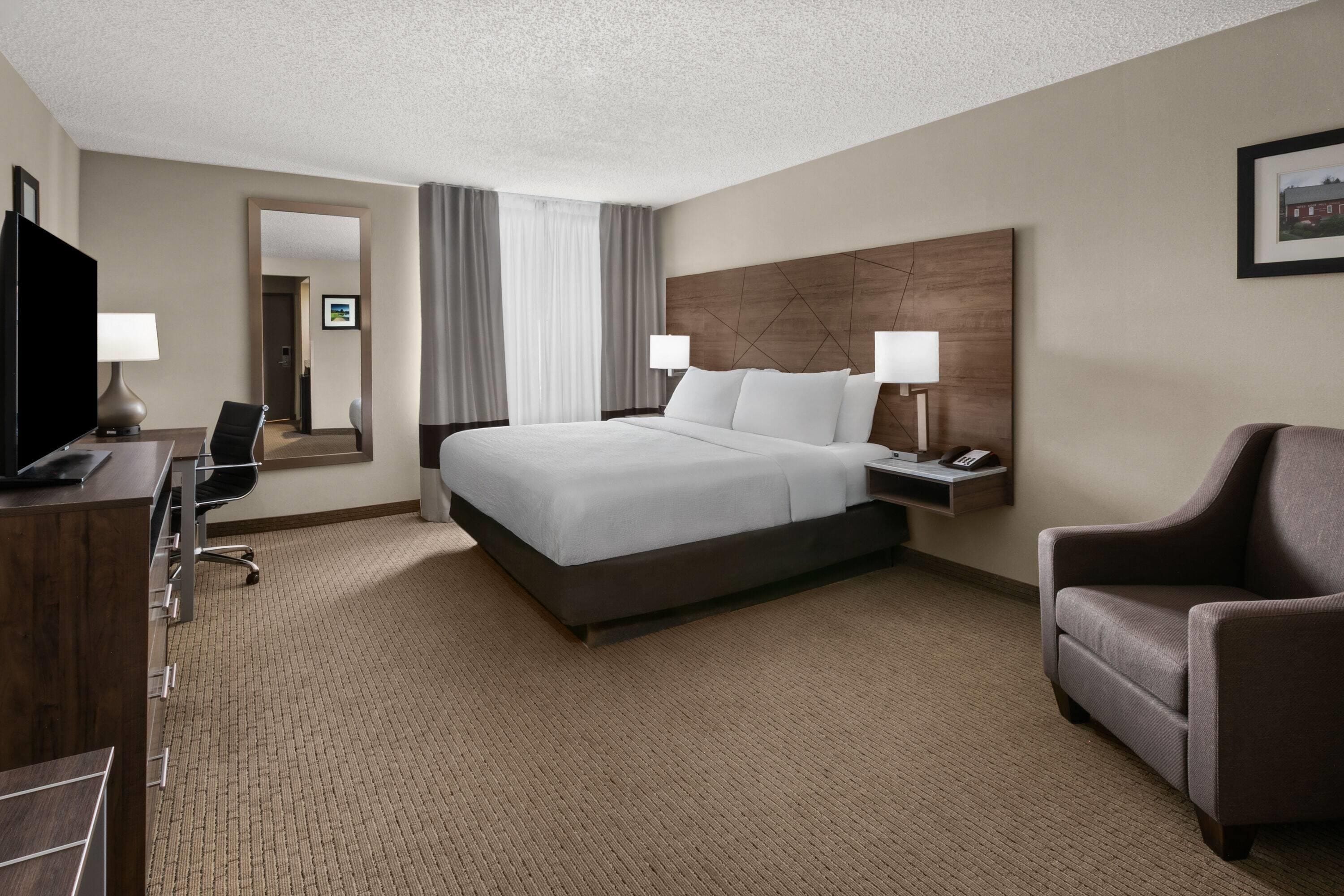 Habitación de hotel con cama, mesita de noche, lámpara, silla, escritorio, televisor y espejo.