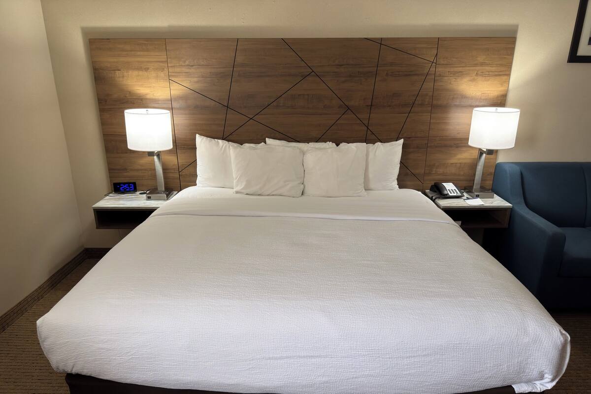 A hotel room with a large bed, two bedside tables, a blue chair, and a wooden headboard.
