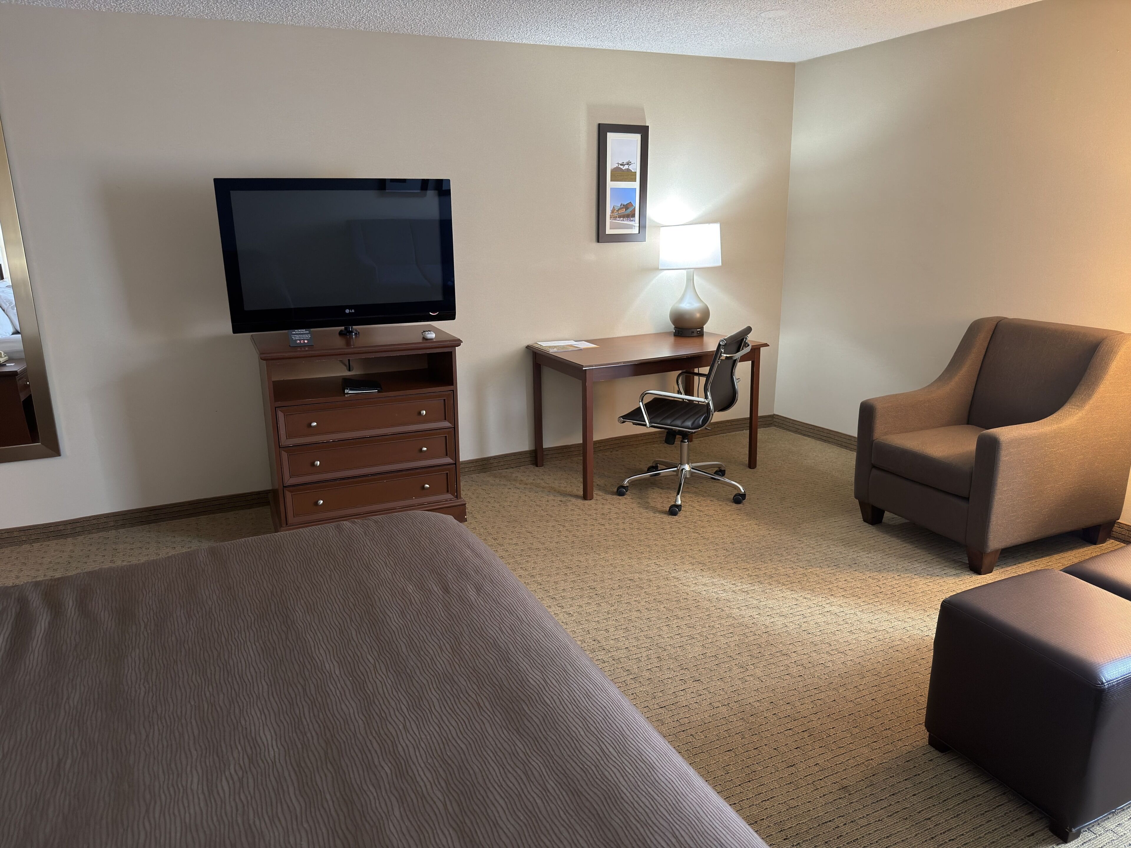 A hotel room with a bed, a desk with a chair, a television, a lamp, and a picture on the wall.