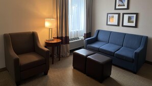 A hotel room with a brown armchair, a blue sofa, two ottomans, a standing lamp, and a window with curtains. - Wingate by Wyndham Utica (Utica)