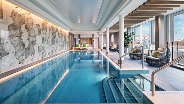 An indoor swimming pool with clear blue water, surrounded by seating areas and large windows offering views of the city. - The Westin Grand Frankfurt (Frankfurt)