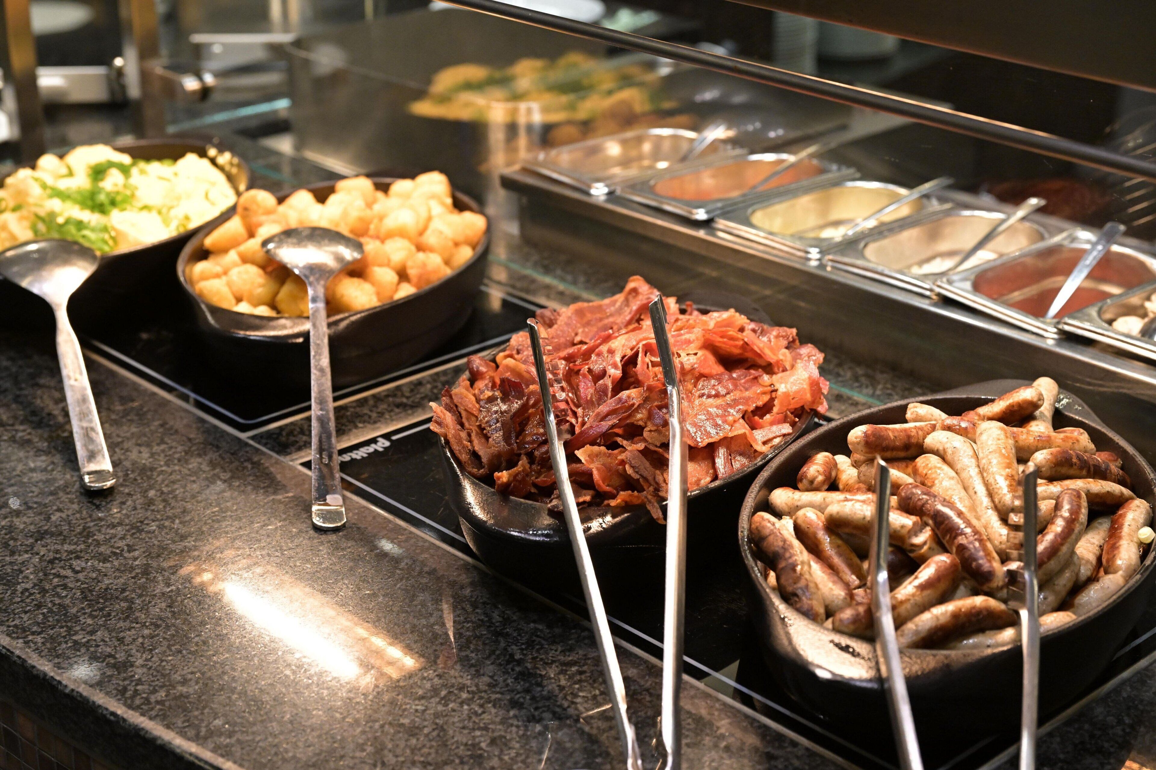 Un bufé con distintos alimentos, incluyendo salchichas, papas y un plato de carne.