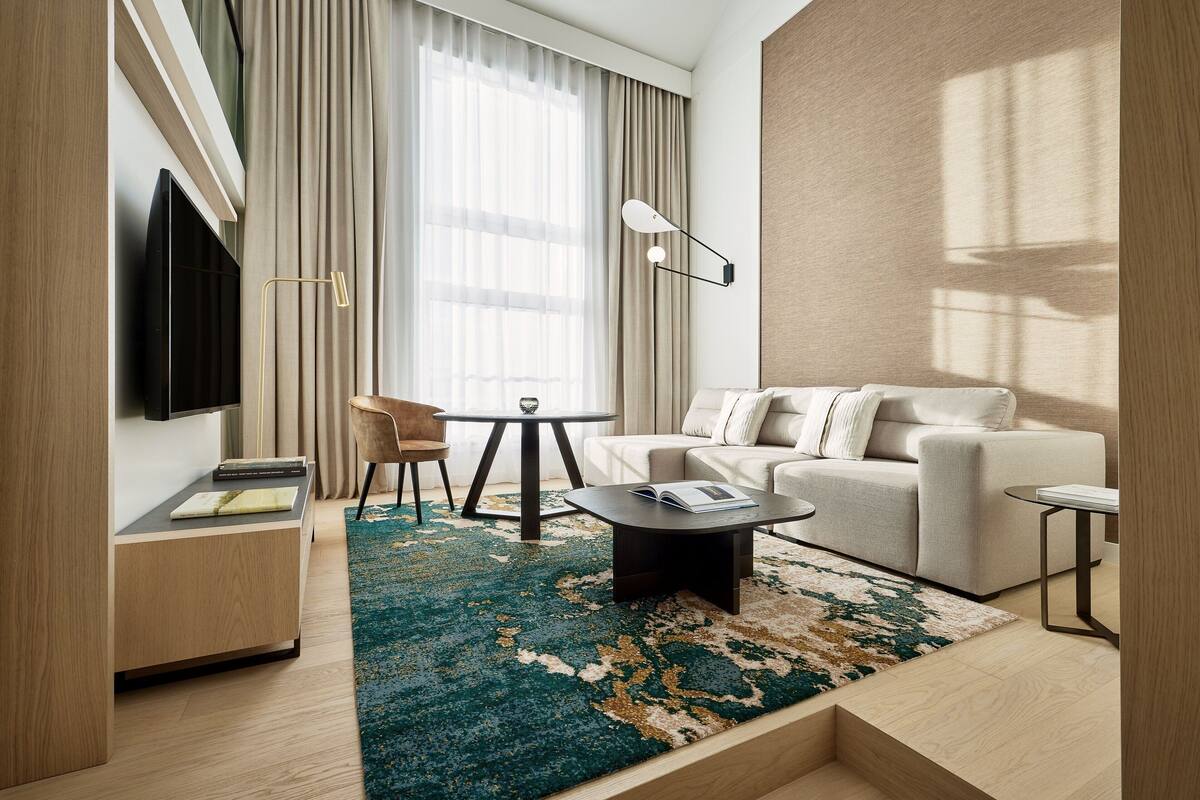 A modern hotel room with a sofa, a coffee table, a TV, a small table with a chair, and a large window with curtains.