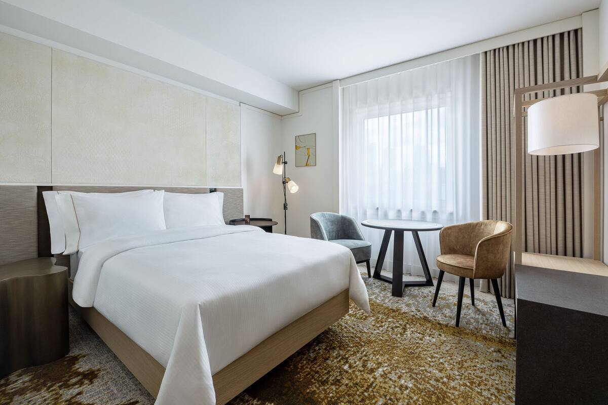 A modern hotel room with a large bed, a desk, a chair, and a small table.