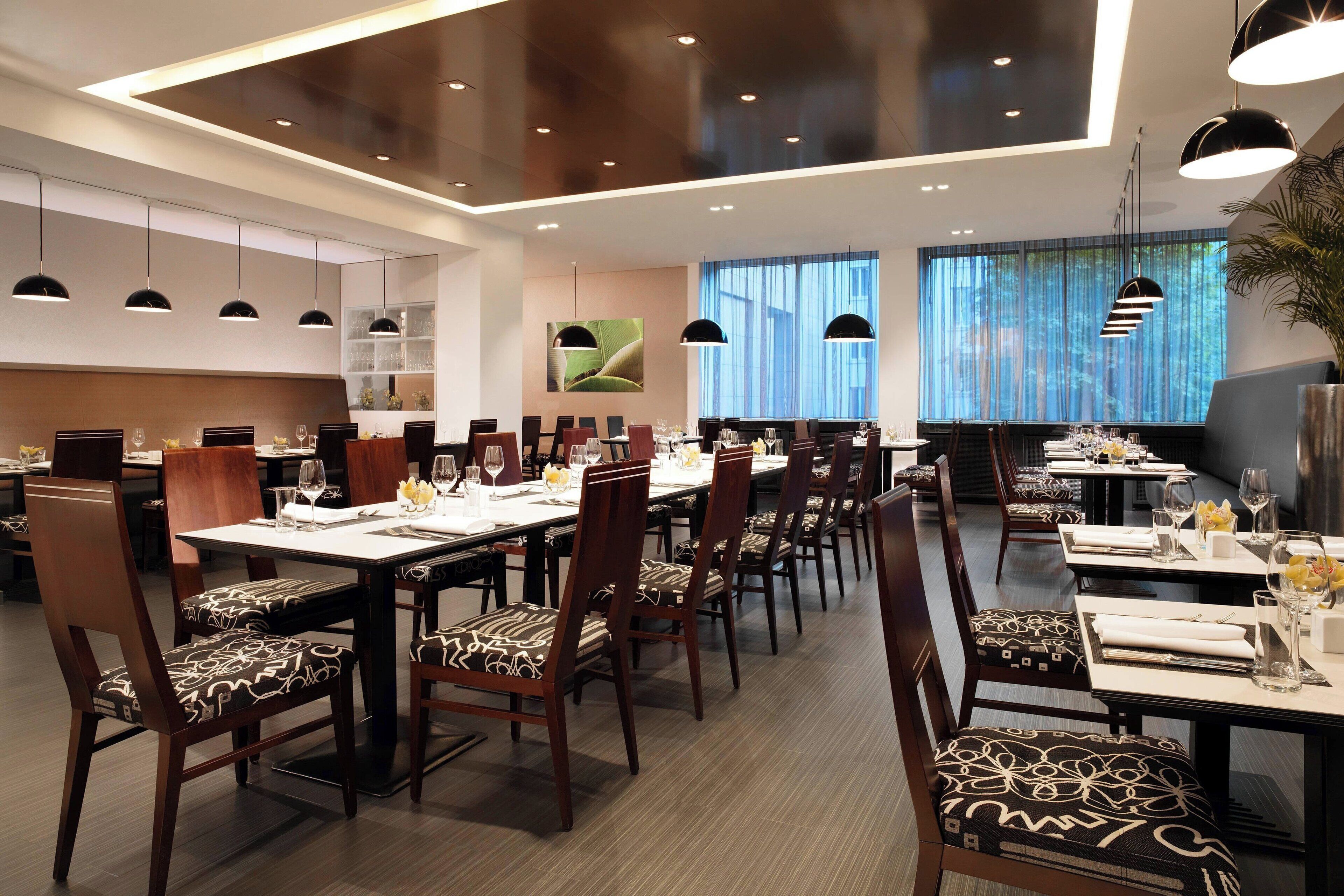 Un comedor moderno con varias mesas y sillas, que destaca por su diseño contemporáneo con lámparas colgantes y un amplio ventanal con cortinas.