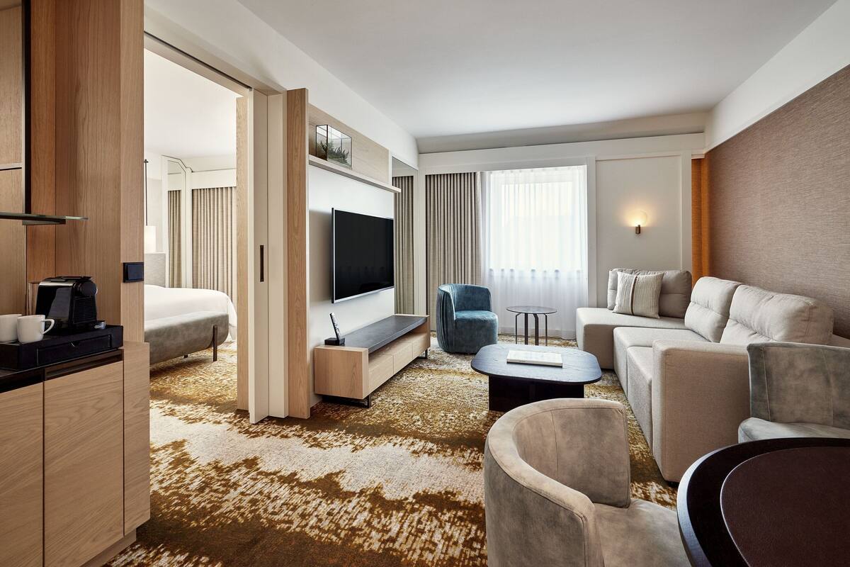 A modern hotel room with a sofa, armchair, and a flat-screen TV mounted on the wall.