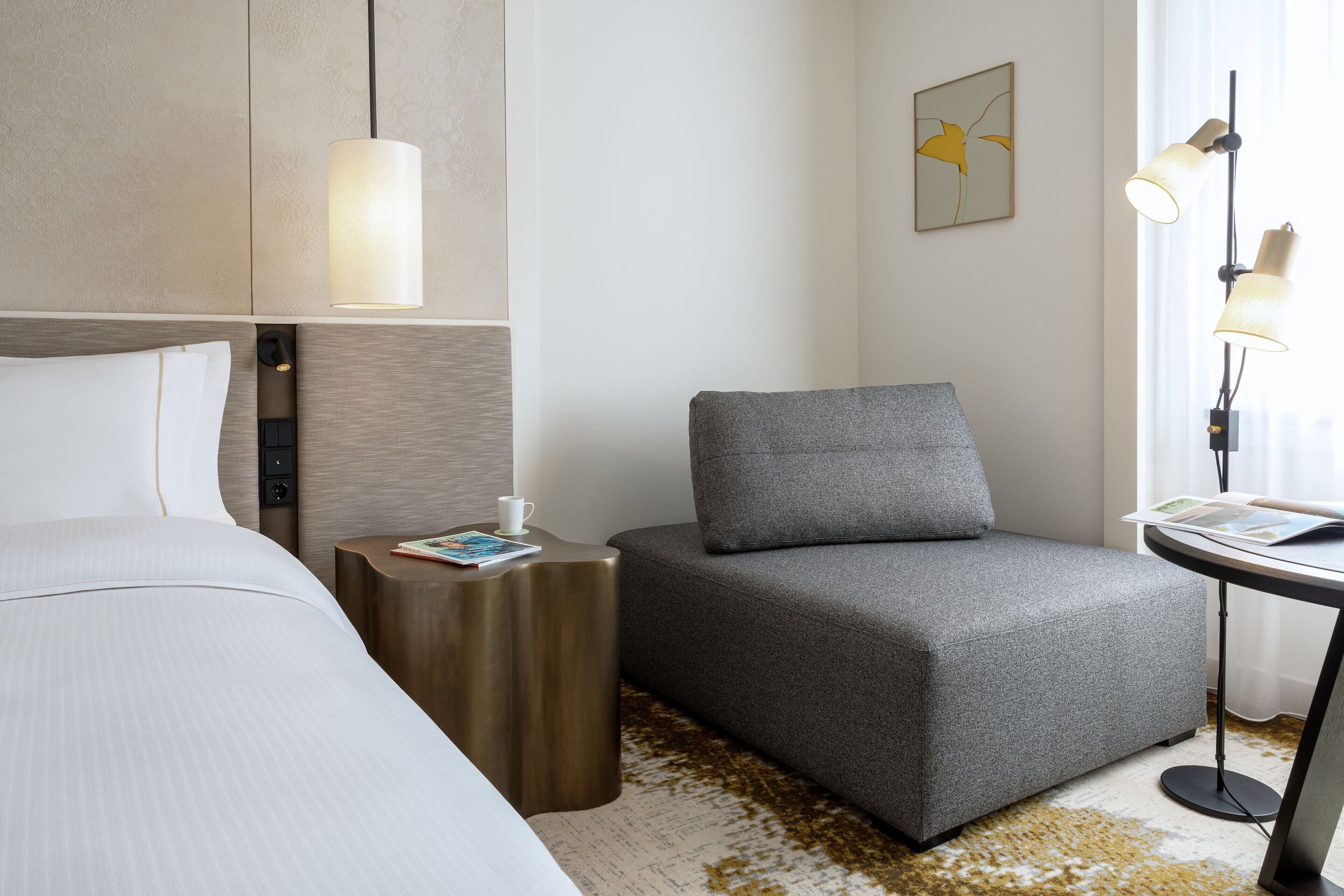 A hotel room with a bed, a gray sofa, a small side table with a book, a lamp, and a painting on the wall.