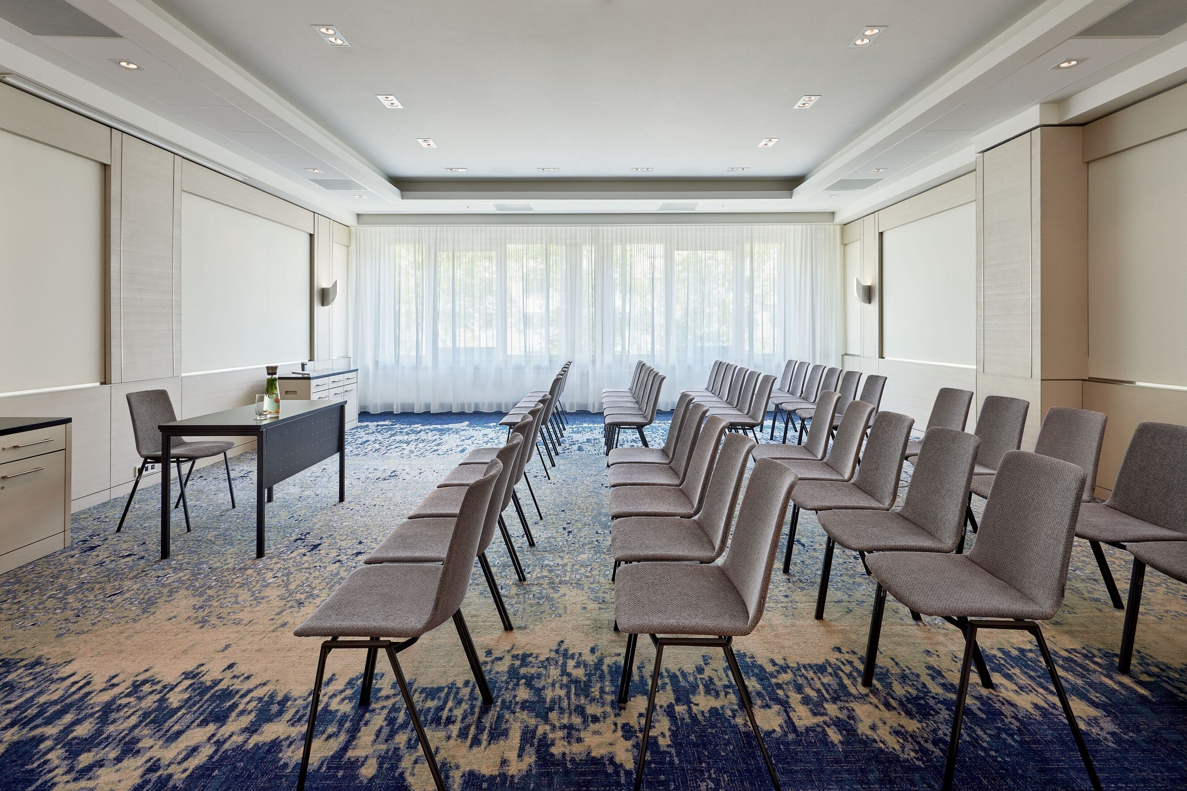 A conference room with a long table and rows of chairs, large windows with sheer curtains, and a patterned carpet.