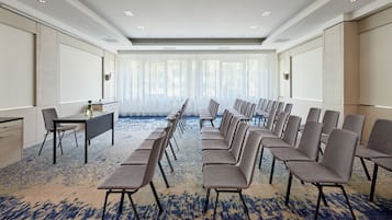 A conference room with a long table and rows of chairs, large windows with sheer curtains, and a patterned carpet.