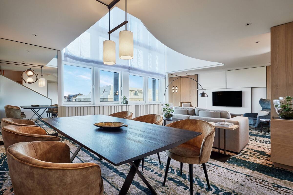 A modern living room with a large dining table, comfortable seating, a flat-screen TV, and large windows offering a city view.
